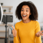 Microinfluencer sonriente creando contenido en video para redes sociales, usando un smartphone y un aro de luz en una sala moderna