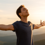 Hombre joven con los brazos abiertos disfrutando del atardecer en la cima de una colina, simbolizando libertad y crecimiento personal.